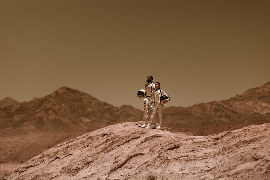 Dwoje astronautów w srebrnych skafandrach stoi na skalistym wzniesieniu przypominającym powierzchnię Marsa.